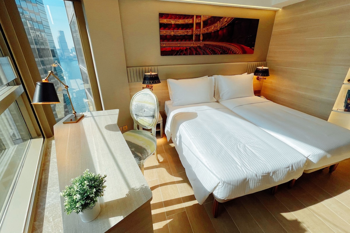 A modern hotel room with a large bed, bedside lamps, a small table with a plant, and a city view through the window.