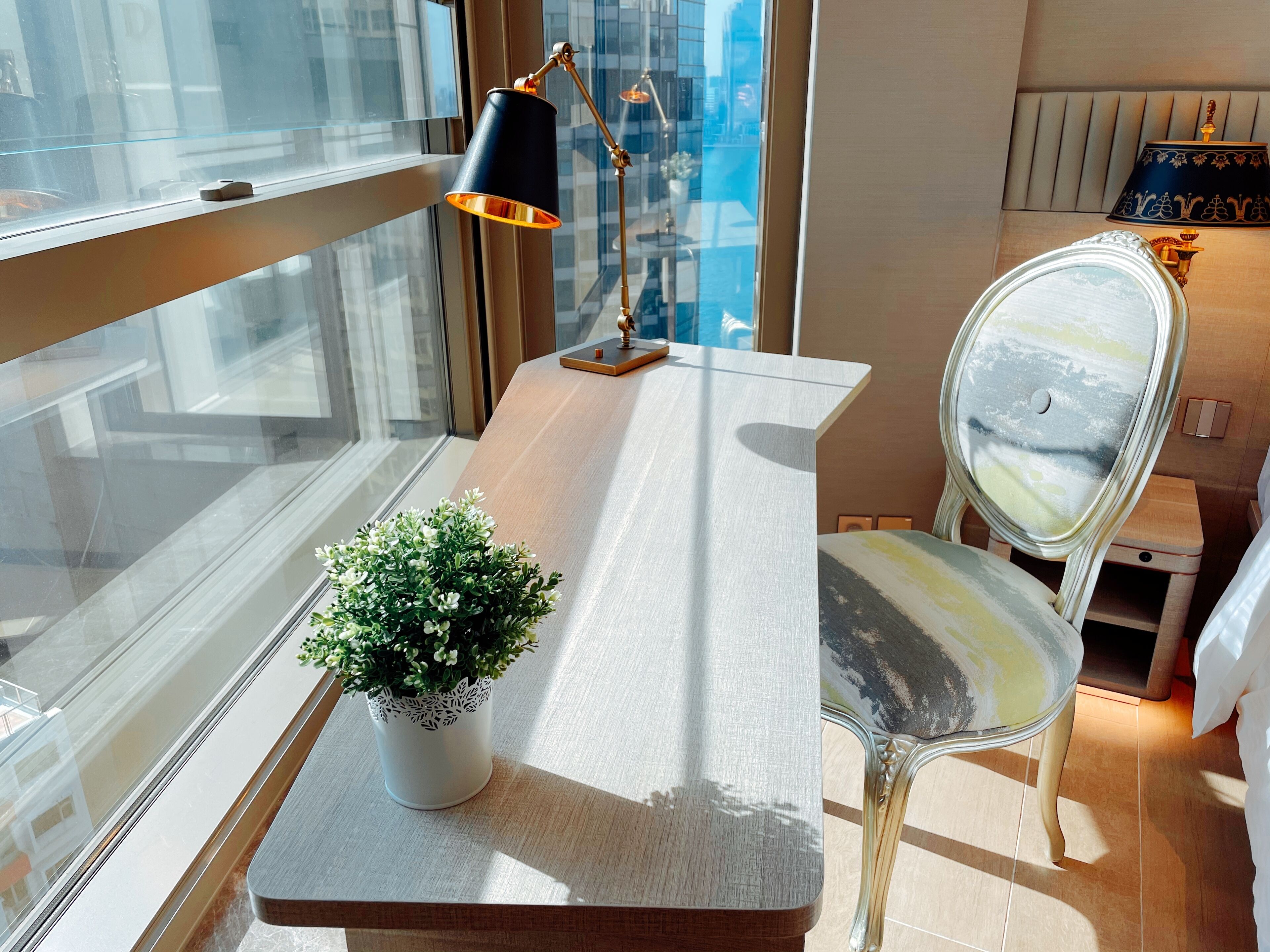 A modern room with a large window, a desk with a potted plant, a chair, and a lamp.