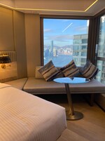 Una habitación de hotel moderna con una cama grande, una zona de estar con cojines, una mesita redonda pequeña y vistas a un paisaje urbano a través de un ventanal amplio.
