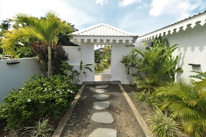 Un sentier de jardin mène à un belvédère blanc surmonté d’un toit en tuiles, entouré d’une végétation luxuriante et de palmiers.