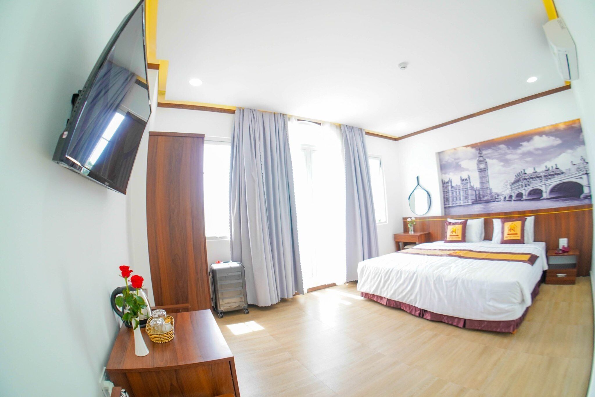 Una habitación de hotel con una cama, un televisor montado en la pared, un escritorio con un florero y un ventanal con cortinas.