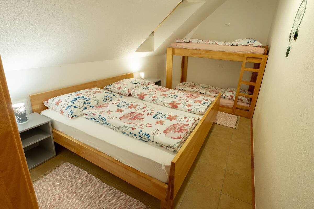 Une petite chambre à coucher accueillante, avec un lit superposé et un lit simple, tous deux dotés de draps à motifs floraux. On y trouve une table de chevet avec une lampe et une petite étagère.