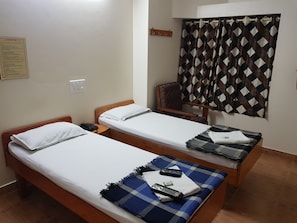 A hotel room with two single beds, a wooden headboard, a small bedside table with a phone, a blanket, and a remote control. - Sri Kumara Lodge (Bengaluru)