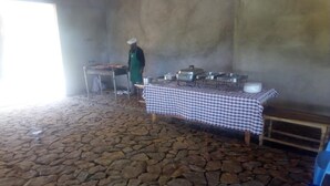 A stone-floored room with a table covered by a checkered tablecloth, a person in a green apron, and various metal containers on the table. - Ruma River Lodge (Ndhiwa)