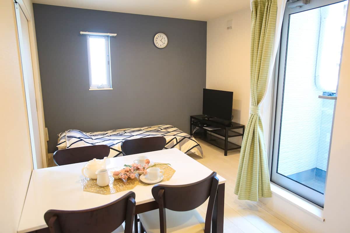 A dining area with a table set for two, a clock on the wall, a television on a stand, and a window with curtains.