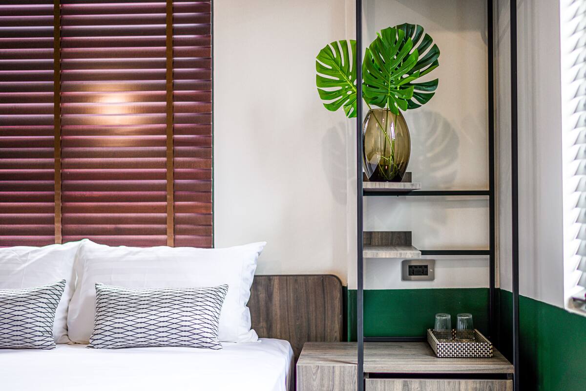 Een hotelkamer met een bed, kussens, een nachtkastje, een plant en een plank met glazen.