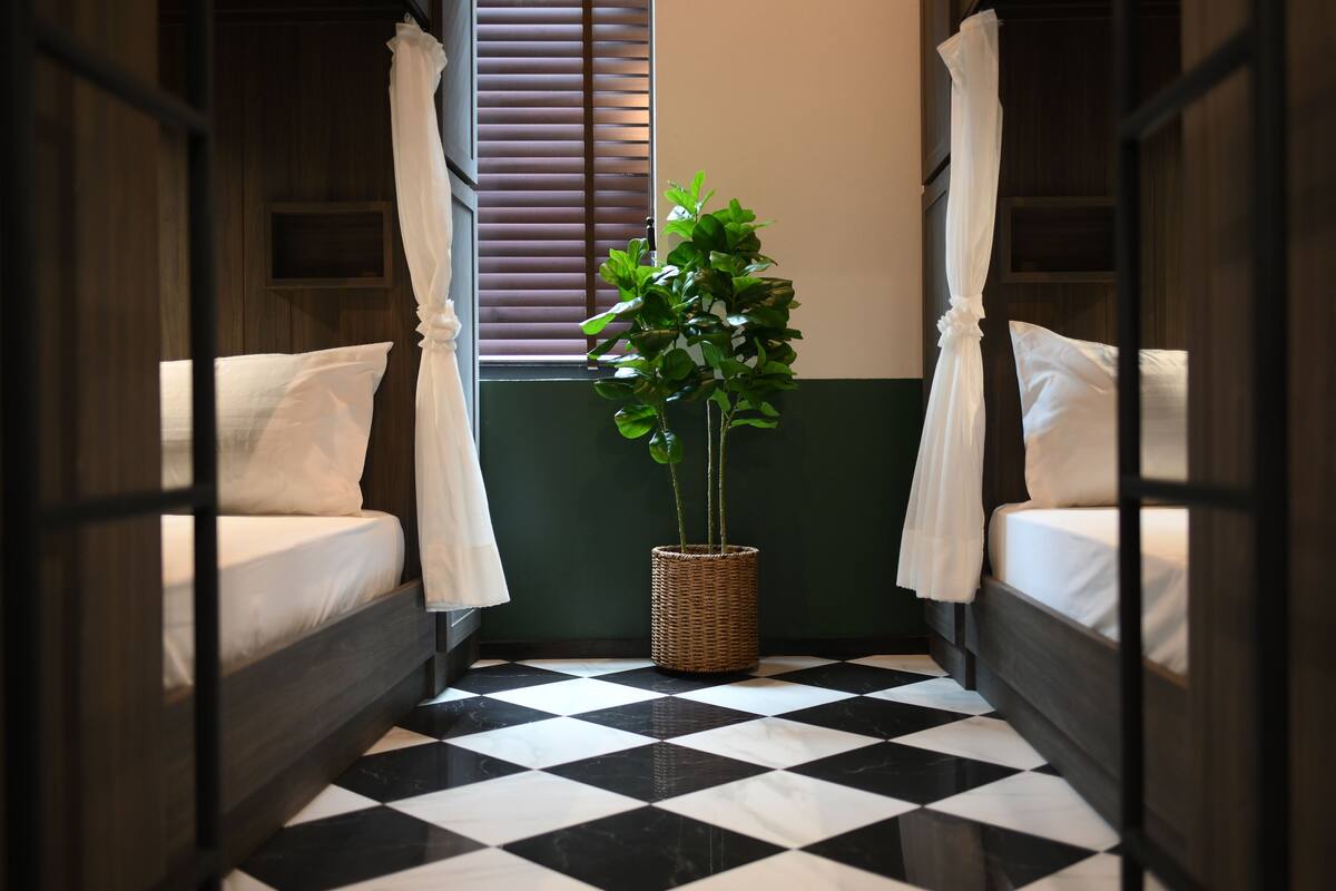 Een kamer met een geruit vloerkleed, twee bedden met witte lakens, een plantenmandje in het midden en een raam met zonwering.