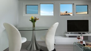 A modern living room with a glass table, two white chairs, a flat-screen TV, a glass coffee table, and a vase with flowers.