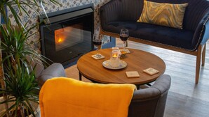 Una sala de estar acogedora con chimenea, un sofá azul oscuro con un cojín amarillo, una mesa de centro redonda de madera y una planta en maceta.