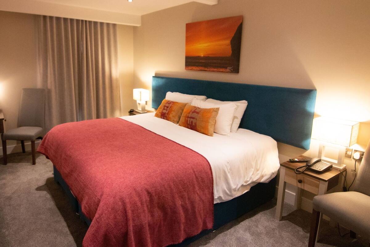 A hotel room with a large bed, a desk with a telephone, a chair, and a painting on the wall.