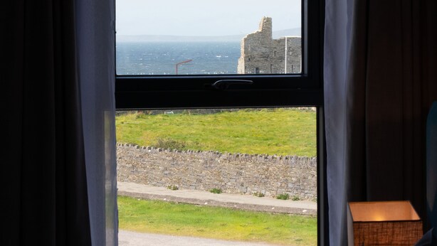 Una ventana con vista a un muro de piedra, un campo cubierto de hierba y un edificio lejano junto al mar.