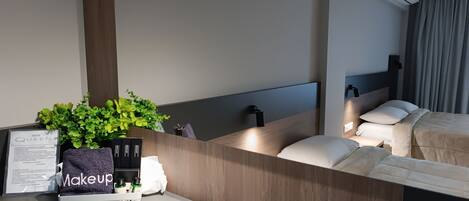 A modern hotel room with a wooden headboard, a bedside table with a makeup bag, a framed menu, and a small plant.