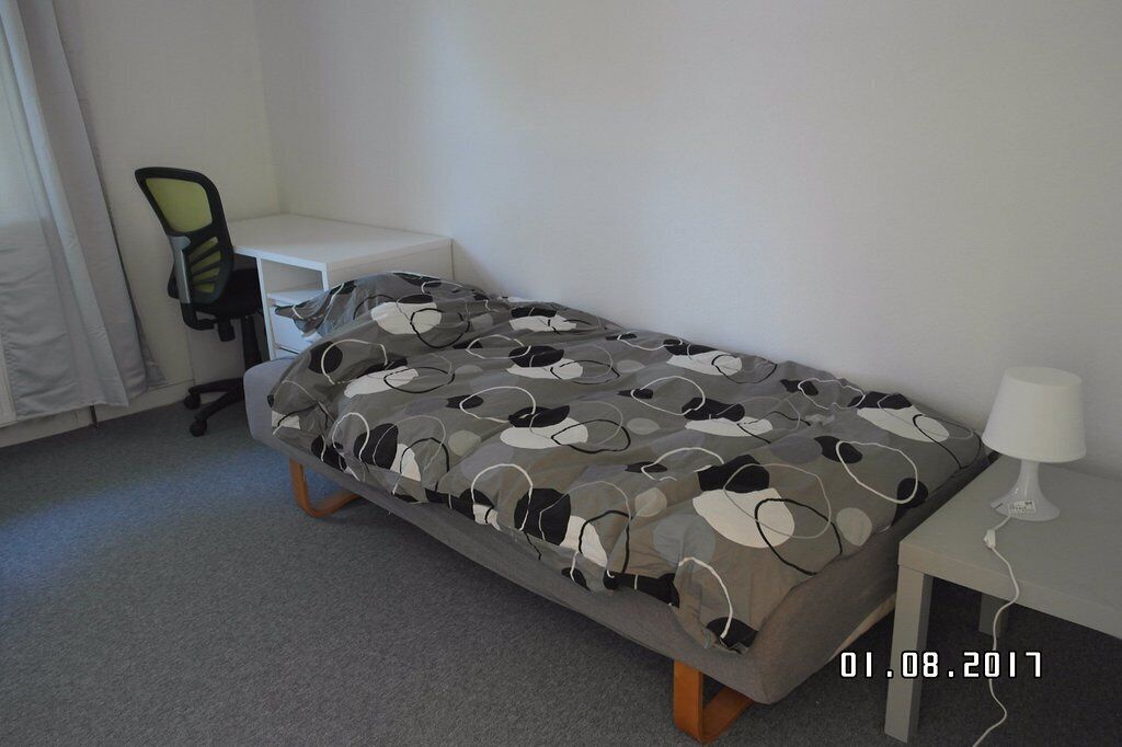 Un lit simple avec une couverture à motifs, une petite table de chevet avec une lampe, une chaise et un bureau blanc.