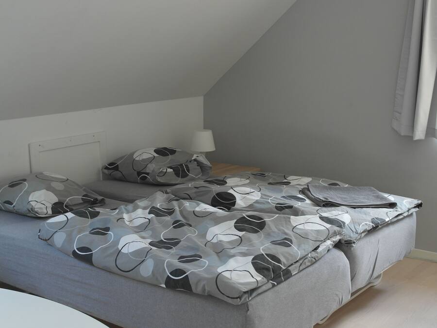 Une chambre à coucher avec un lit, des oreillers, une table de chevet, une lampe et une fenêtre avec des rideaux.