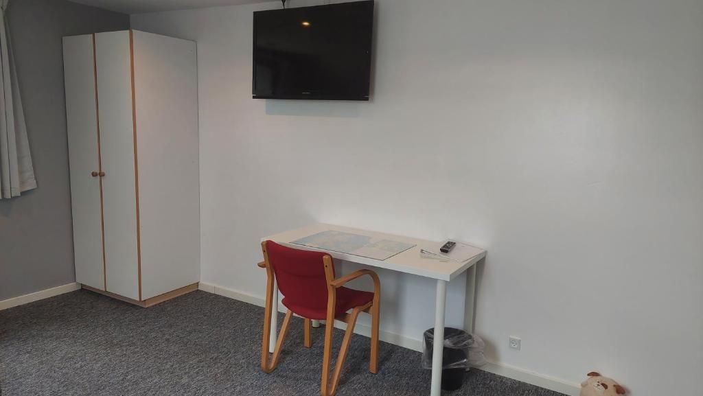 Een kamer met een wit bureau, een rode stoel, een kledingkast en een aan de muur bevestigde televisie.