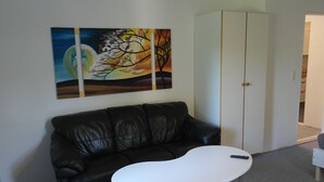 A living room with a black leather couch, a white coffee table, and a painting of a tree against a sunset. - Ringslund B&B (Galten)