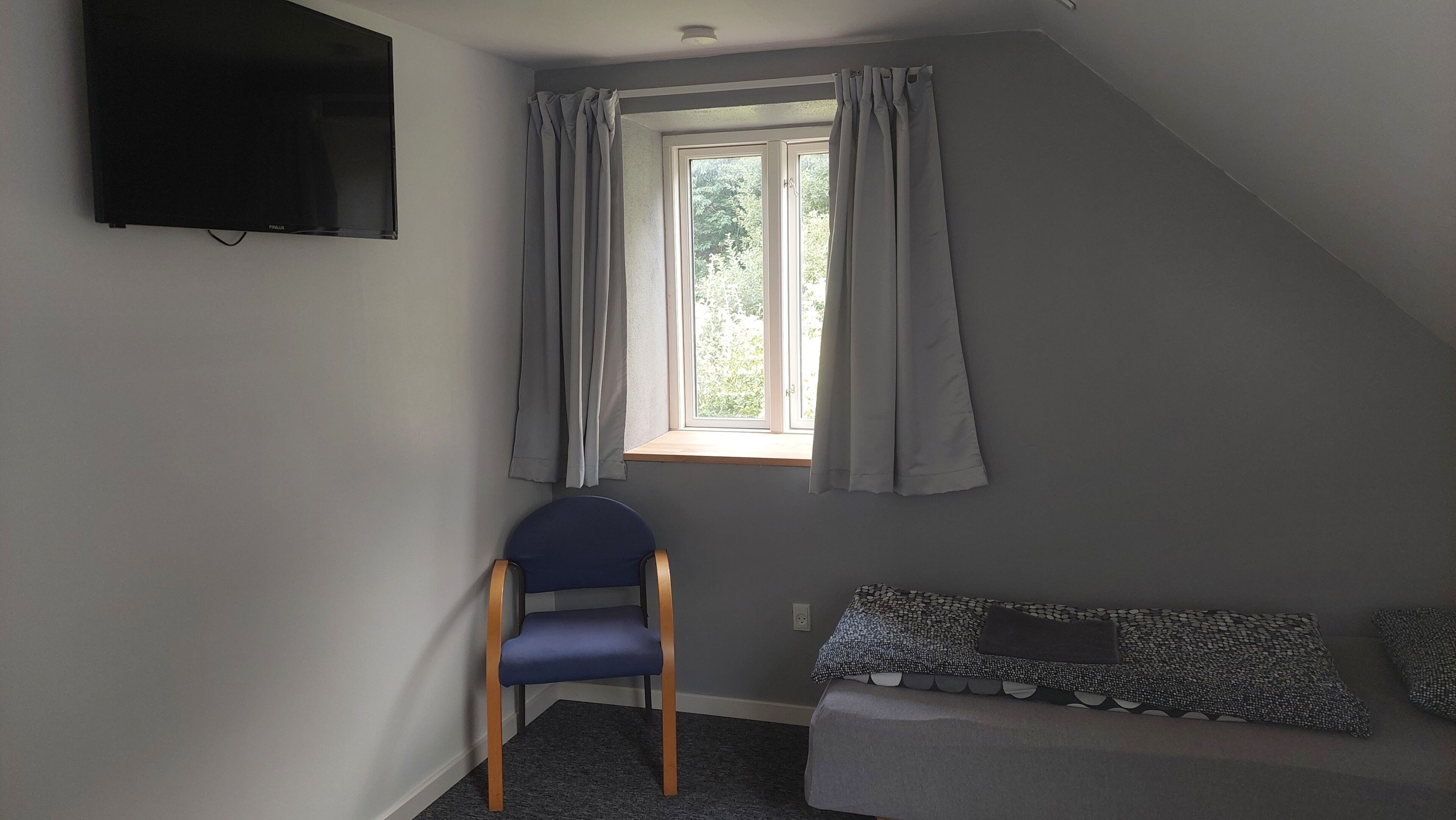 Una habitación pequeña, tenuemente iluminada, con una sola silla, un banco y una ventana con cortinas.