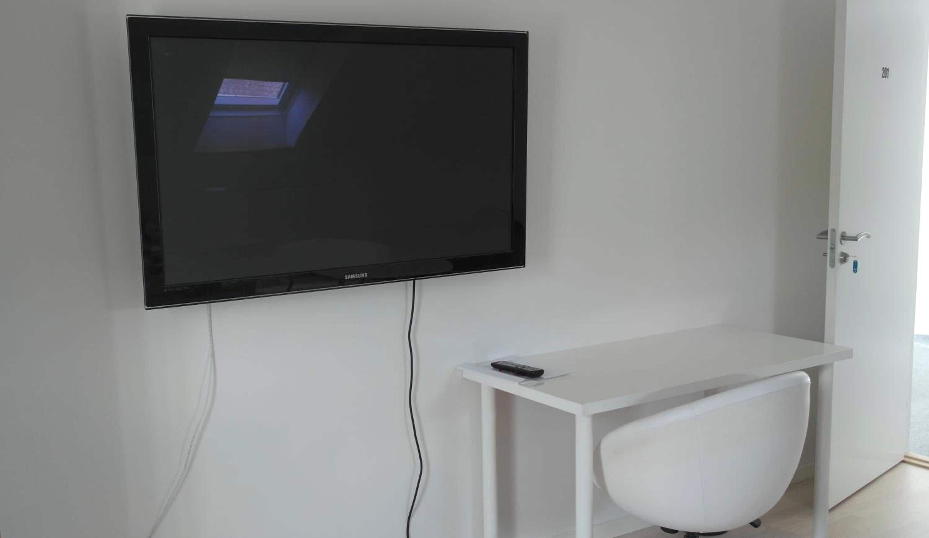 Un televisor de pantalla plana montado en una pared blanca, un escritorio blanco con un control remoto y una silla blanca.