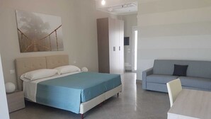 A modern hotel room with a bed, a sofa, a dining table, and a large abstract painting on the wall. - Sand Hotel (Botricello)