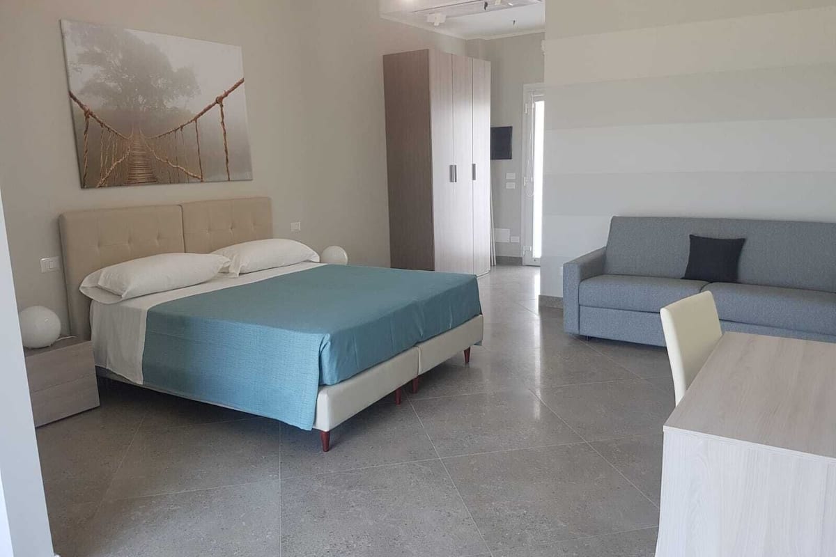 A modern hotel room with a bed, a sofa, a dining table, and a large abstract painting on the wall.