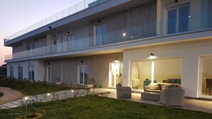 A modern two-story building with a flat roof, large windows, and a well-lit outdoor seating area with wicker furniture. - Sand Hotel (Botricello)