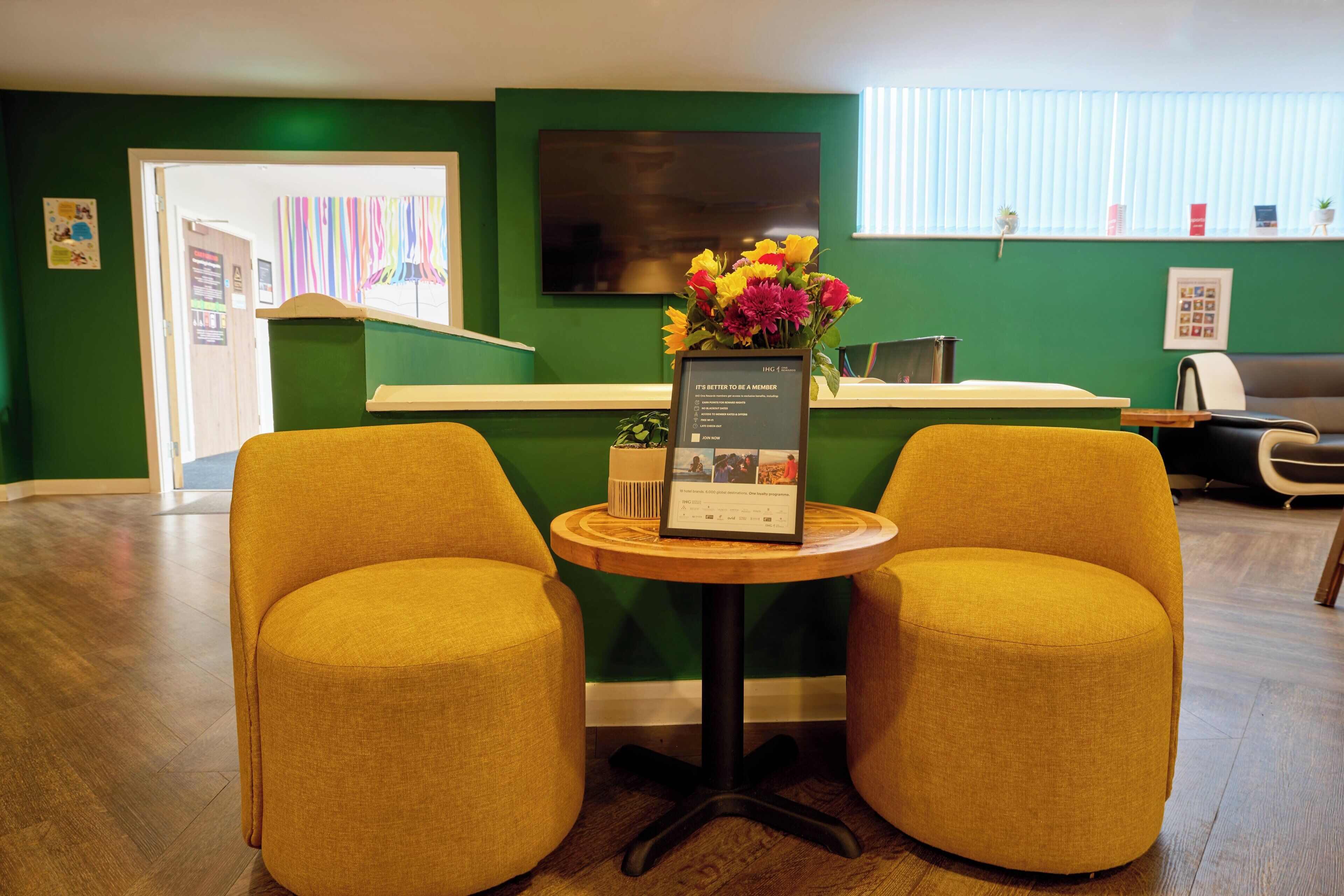 A reception area with a green wall, a flat-screen TV, a wooden table with a framed certificate, and two yellow armchairs.