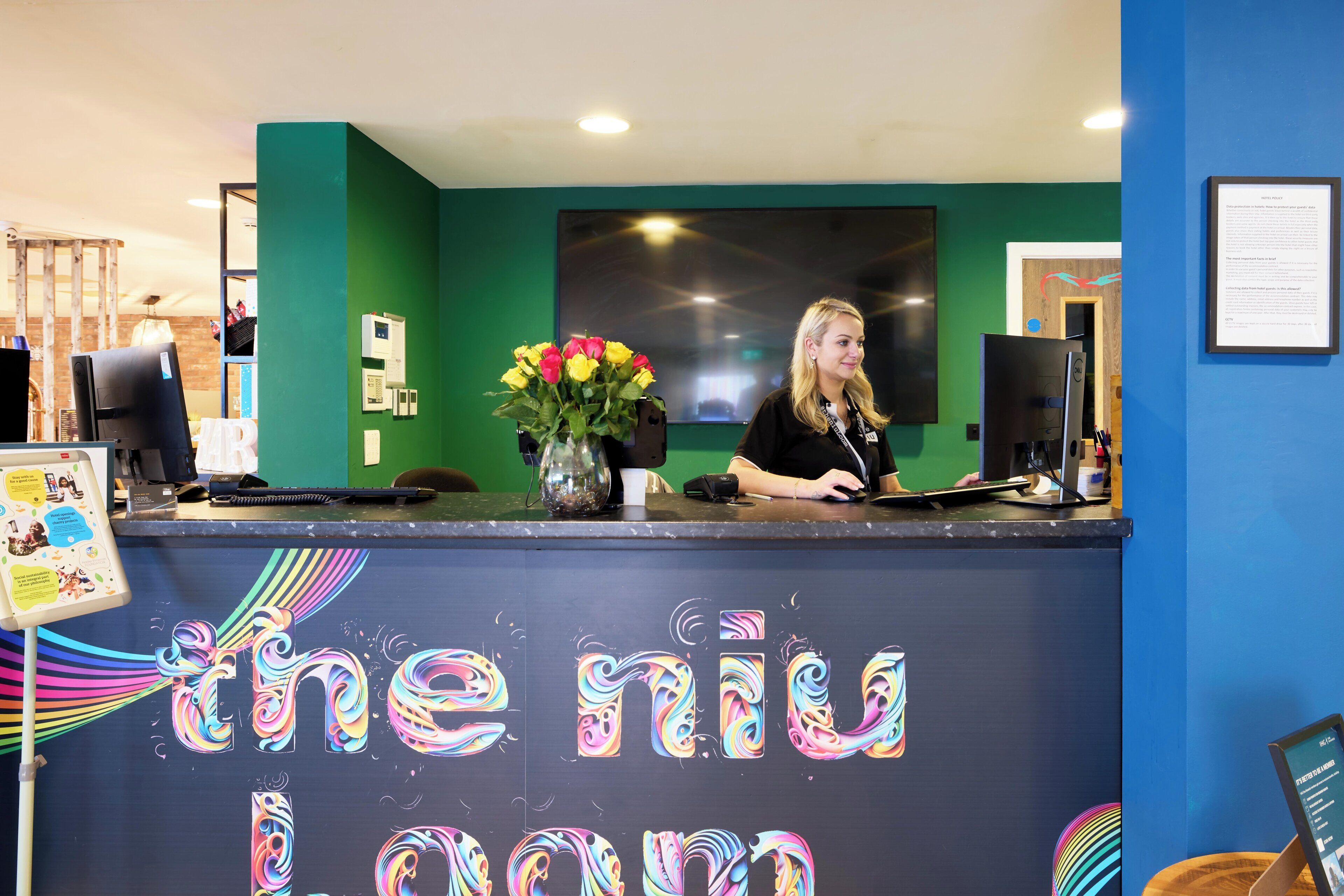 A reception desk with a colorful, swirling design on the wall, a vase with flowers, and a person seated behind the desk.