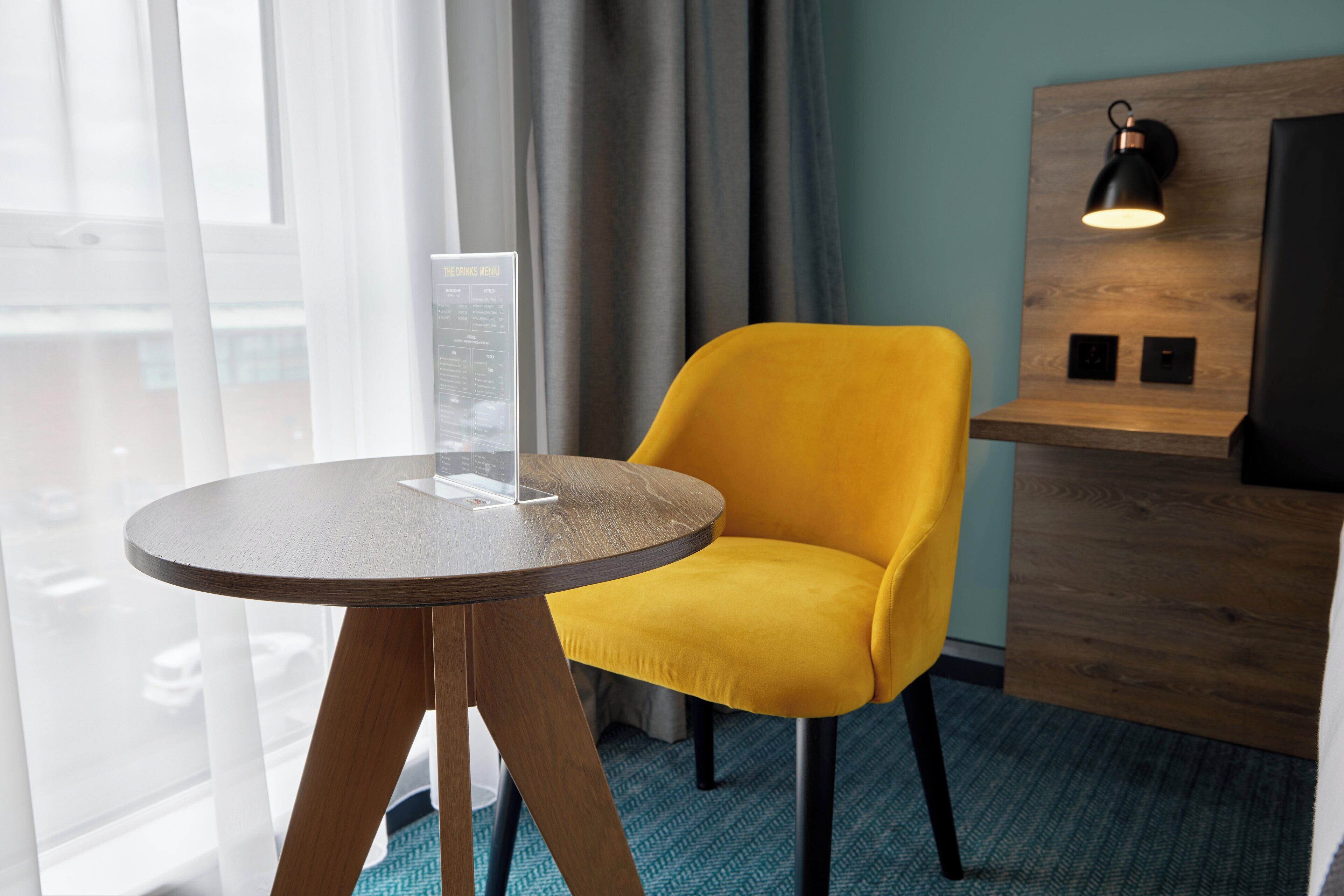 A hotel room with a round wooden table, a yellow chair, a wooden headboard with a lamp, and a view through large windows.