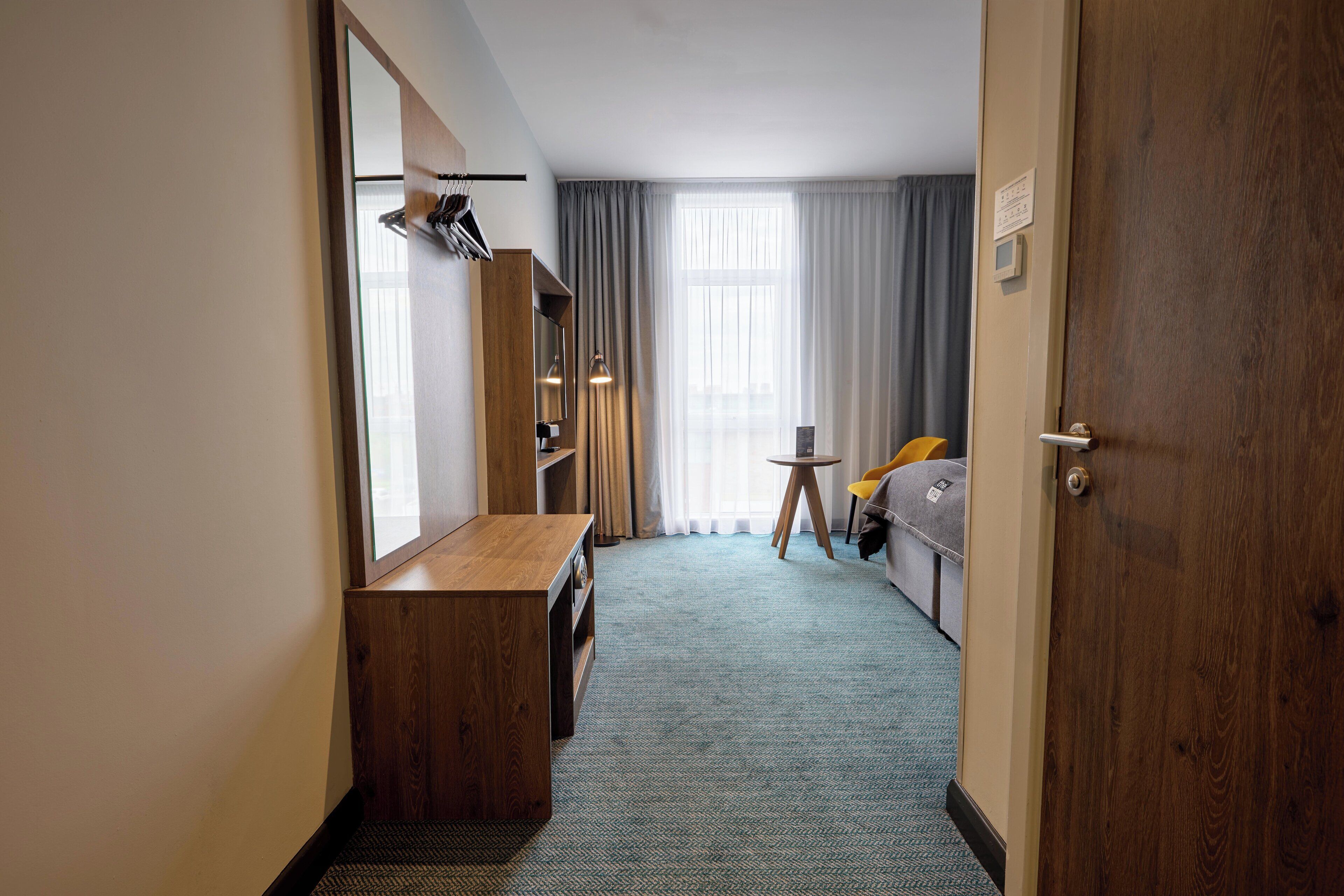 A hotel room with a wooden desk, a bed, a small table with a chair, and a large window with curtains.