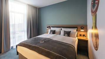 Una habitación de hotel con cama, cortinas, mesita de noche, lámpara y un adorno en la pared.
