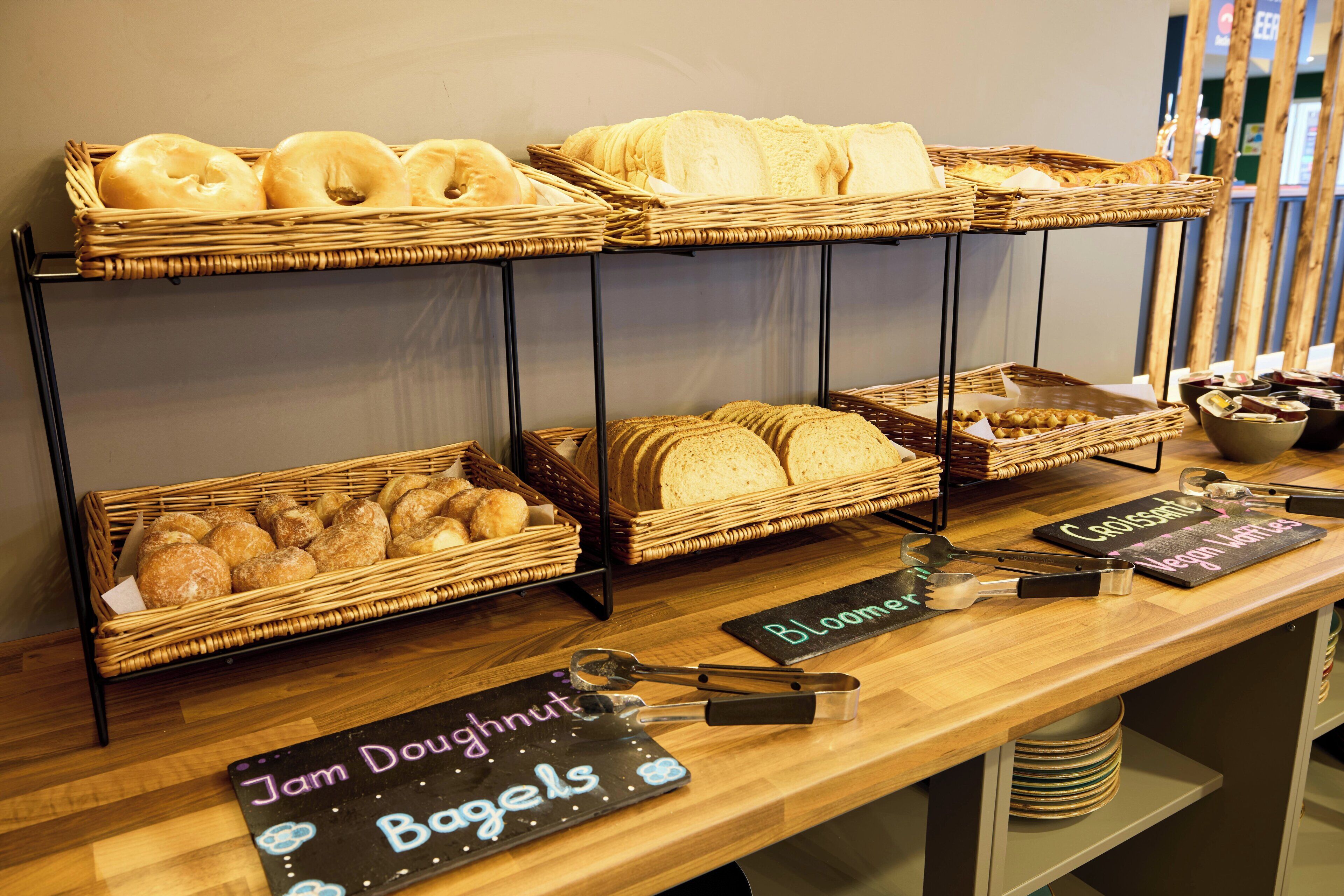 Una mesa de buffet con diversas opciones de productos horneados, como donas y pan, y etiquetas que identifican diferentes tipos de pastelería.