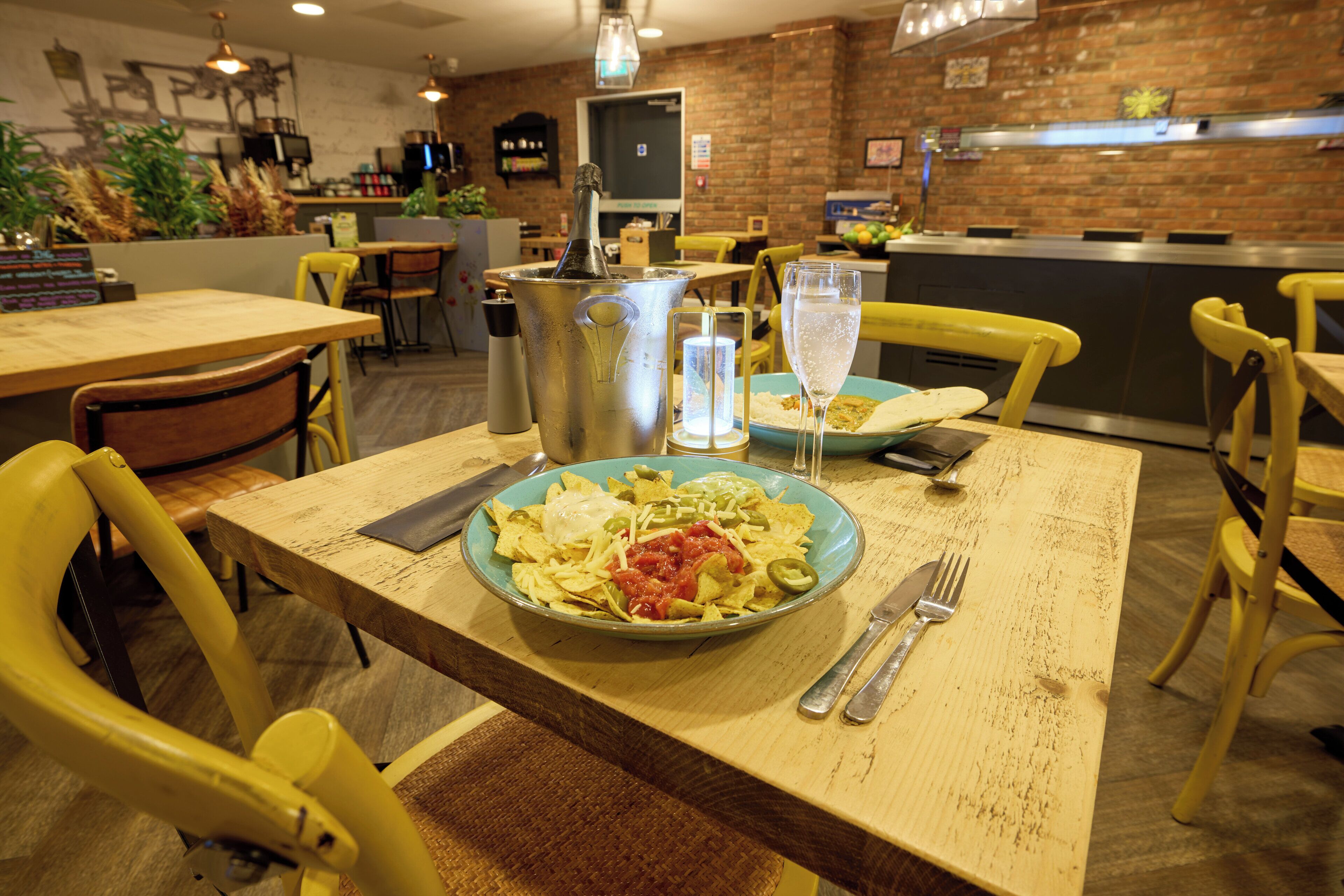 Interior de restaurante com parede de tijolos, mesas de madeira, cadeiras amarelas e um prato de nachos azuis.
