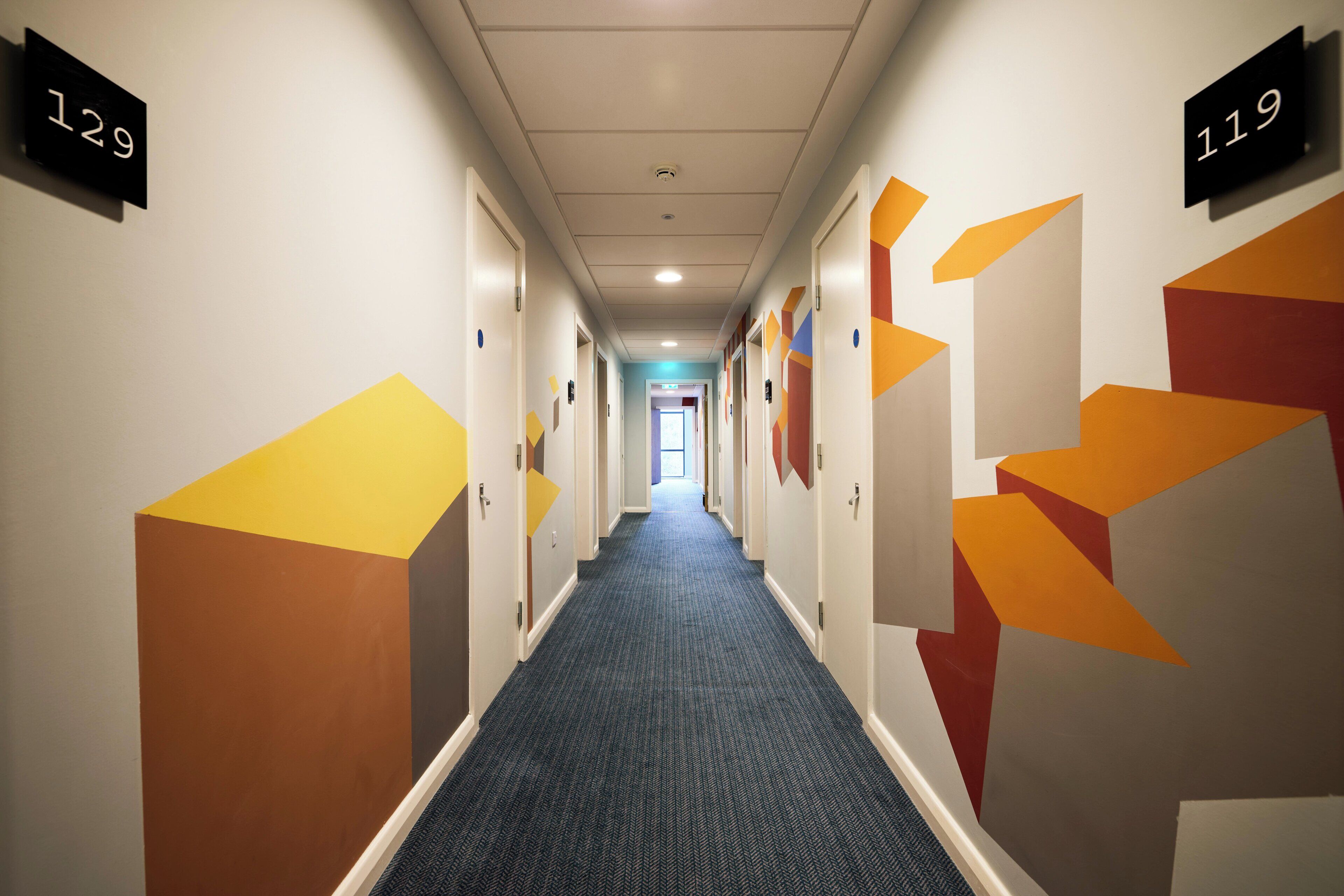 A hallway with doors numbered 129 and 119, carpeted floor, and geometric wall decorations.
