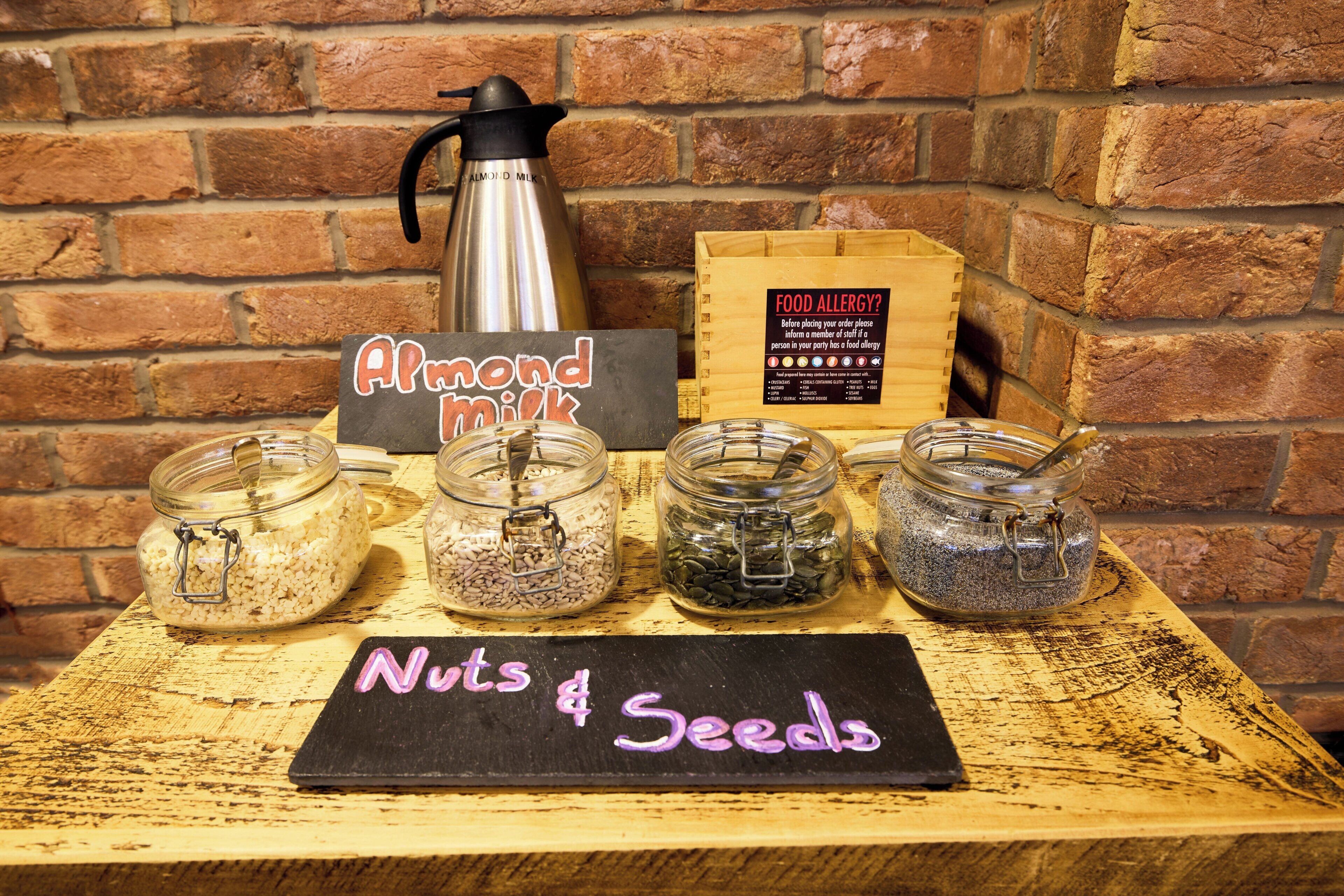 A wooden table with jars labeled "Almond Milk" and "Nuts & Seeds," a sign for food allergies, and a coffee maker.