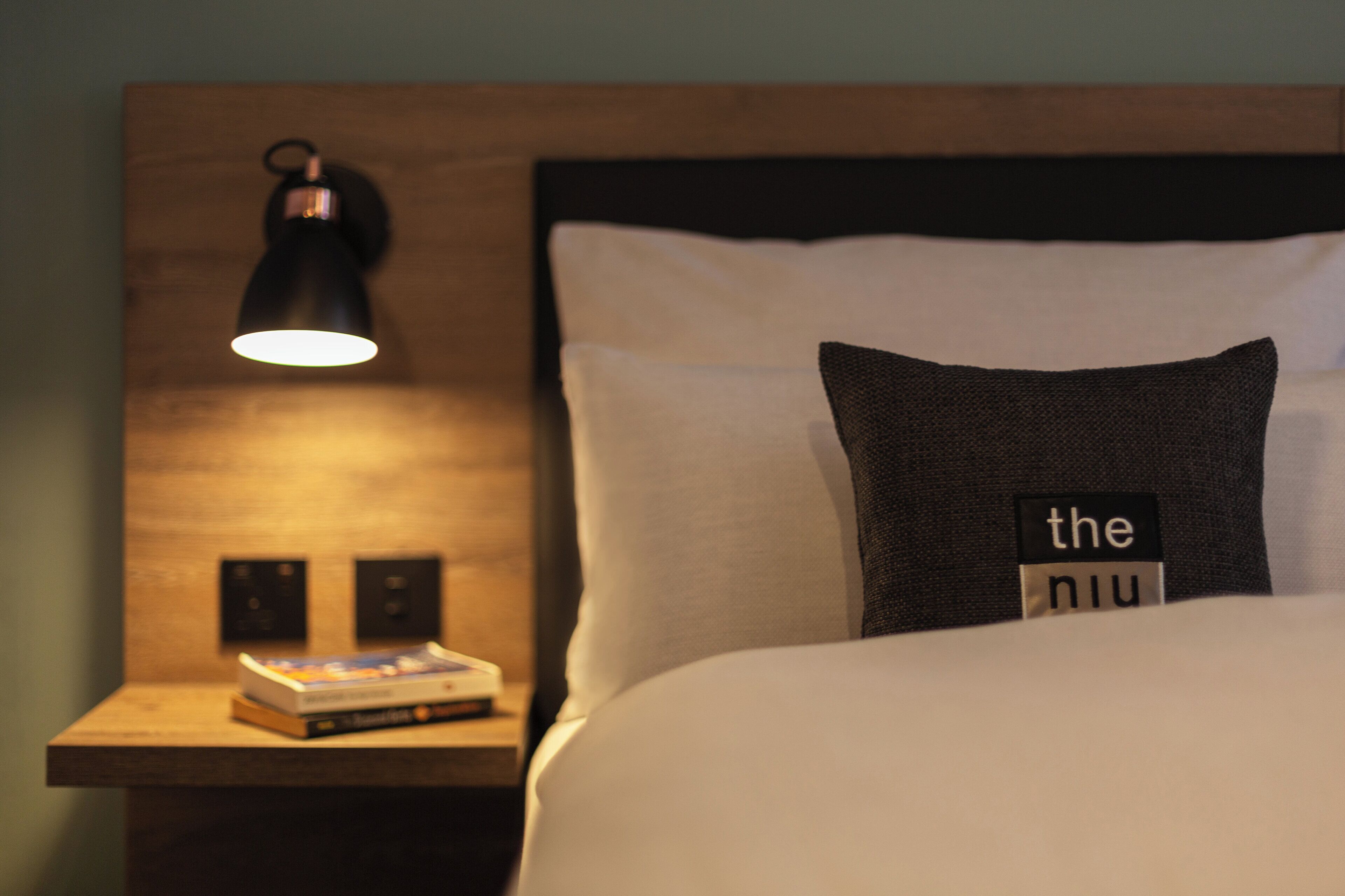 A neatly made bed with a pillow labeled "the niu" and a bedside table with a book and a lamp.