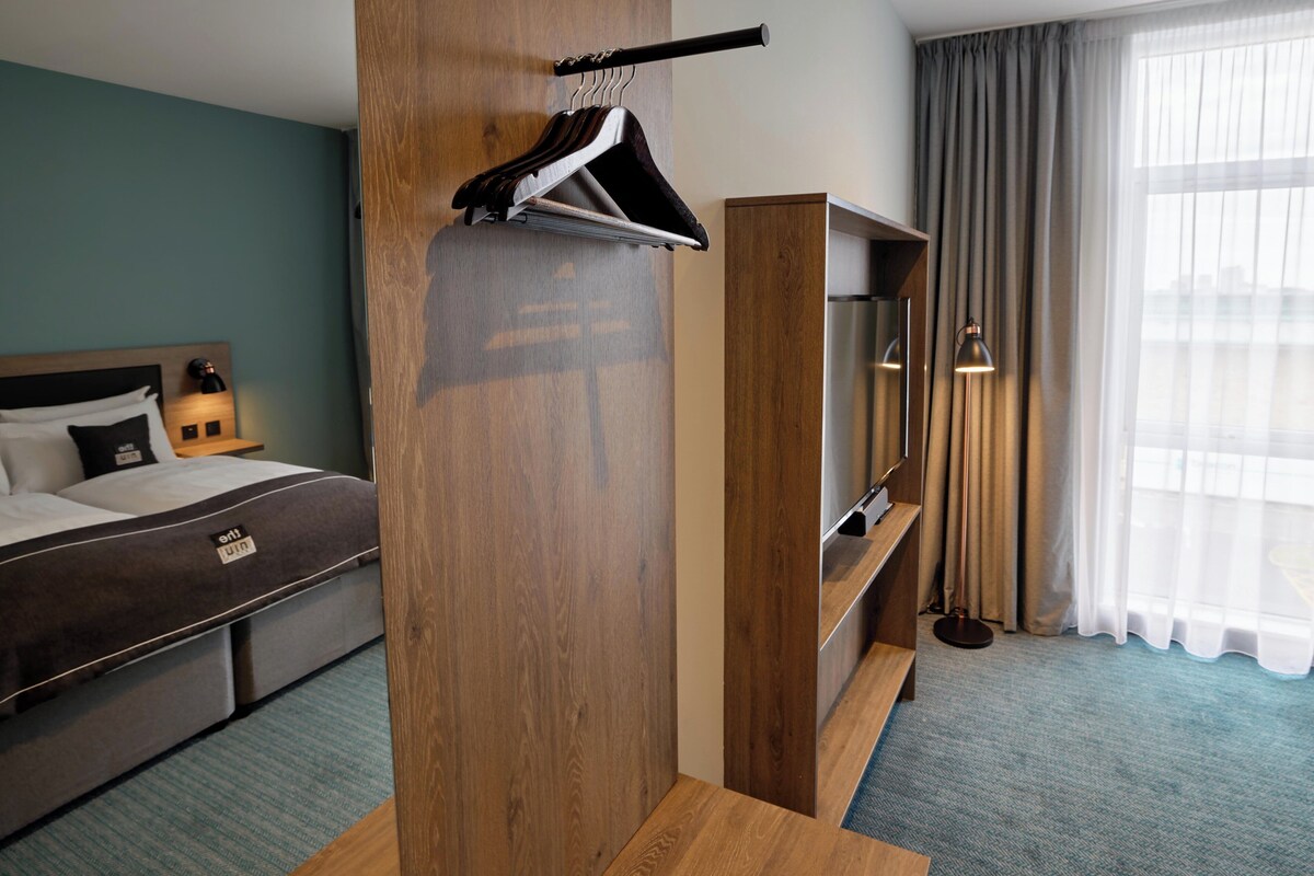 A hotel room with a bed, a wooden wardrobe, a TV, and a window with curtains.