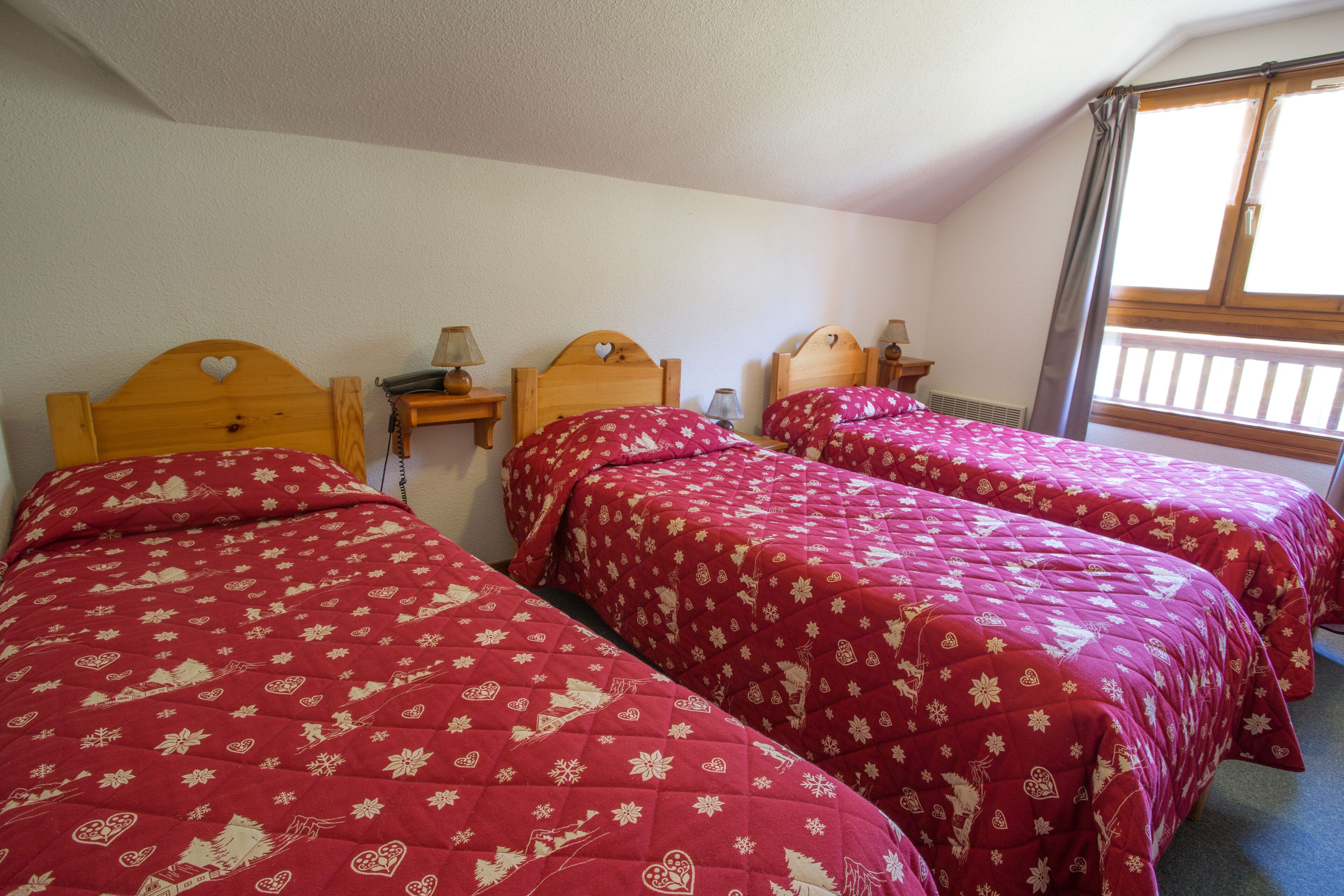 Ein Zimmer mit zwei hölzernen Betten, ausgestattet mit roter floraler Bettwäsche, Holz-Kopfteilen, Nachttischen inklusive Lampen und einem Fenster mit Blick auf einen Balkon.