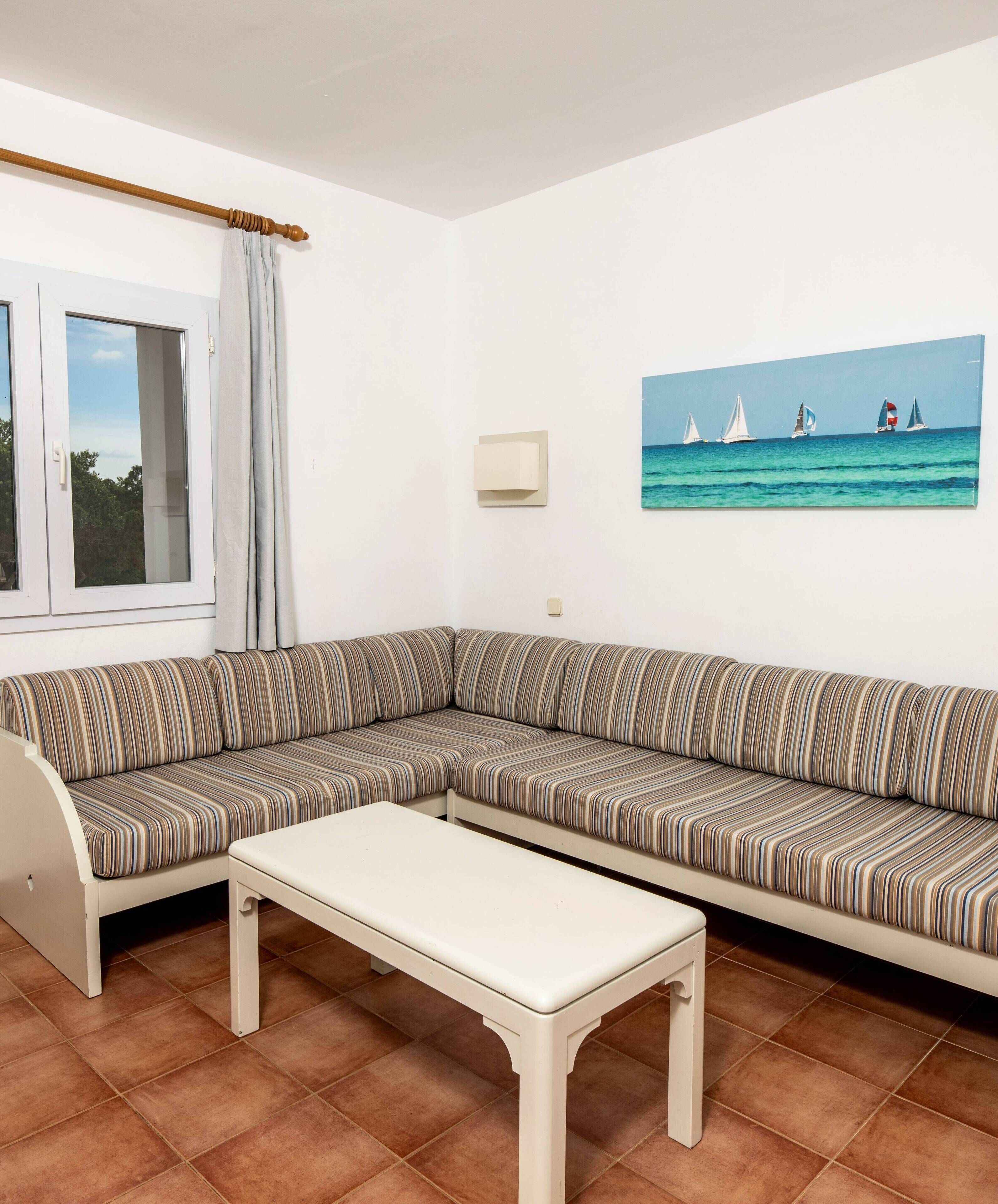 A living room with a striped sofa, a white coffee table, a window with curtains, and a painting of sailboats on the wall.