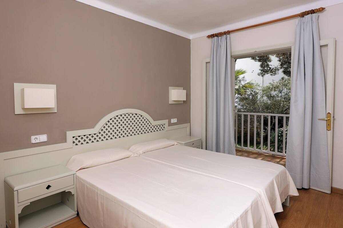 A bedroom with a bed, bedside tables, a nightstand, a window with curtains, and a view of greenery outside.