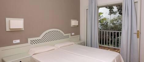 A bedroom with a bed, bedside tables, a nightstand, a window with curtains, and a view of greenery outside.