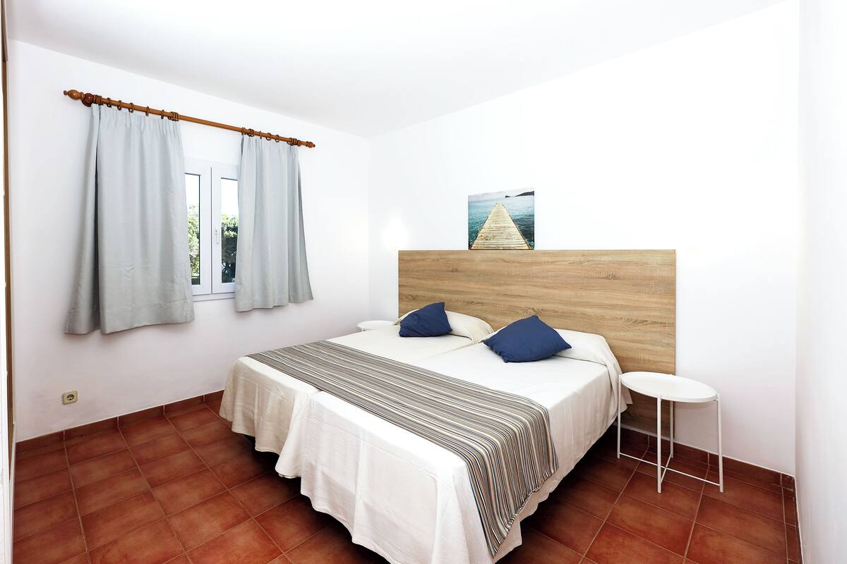 A bedroom with a bed, curtains, a headboard, a bedside table, and a wall-mounted picture.
