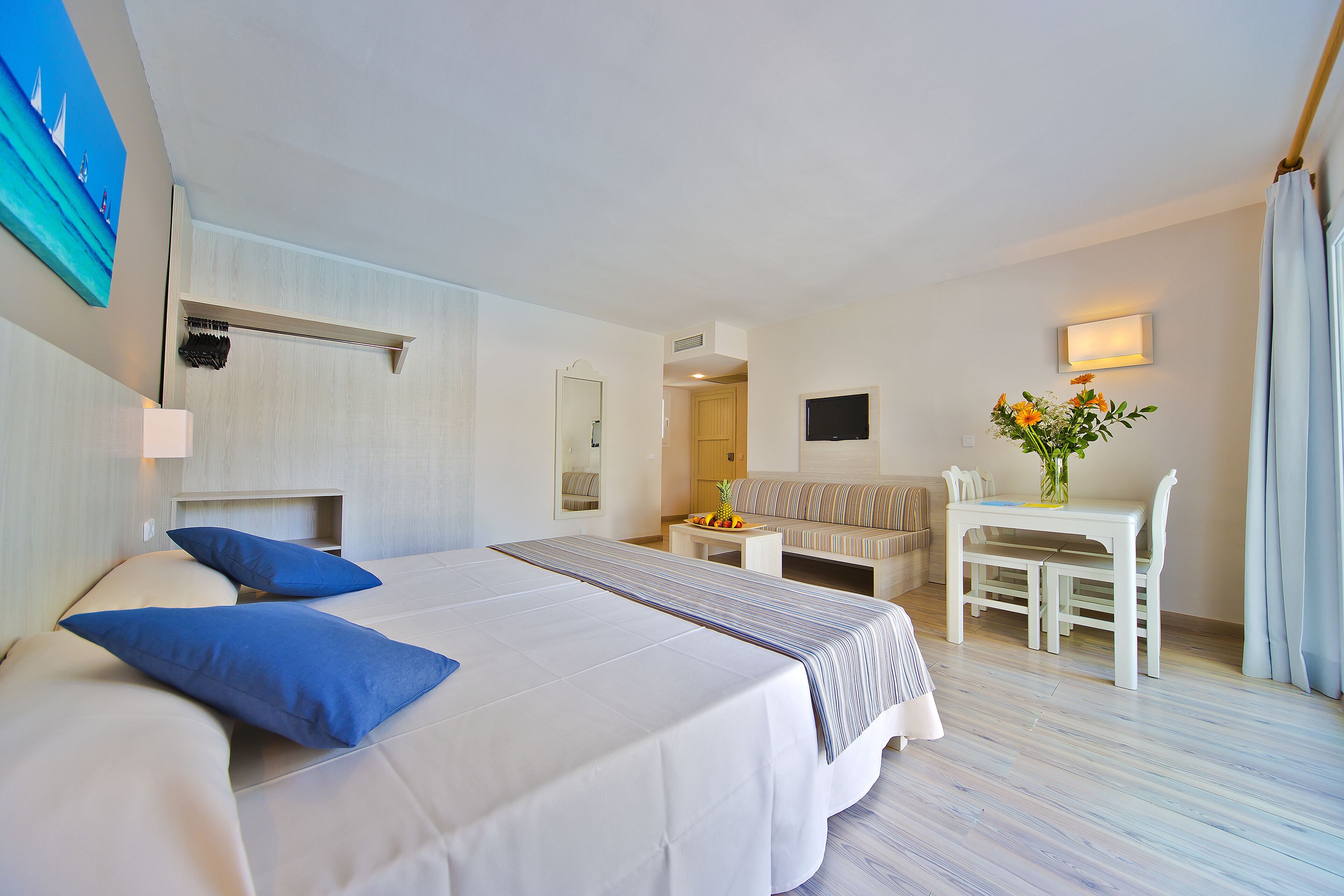 A hotel room with a large bed, a desk, a chair, a small table with fruit, and a painting on the wall.