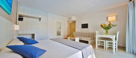 A hotel room with a large bed, a desk, a chair, a small table with fruit, and a painting on the wall.