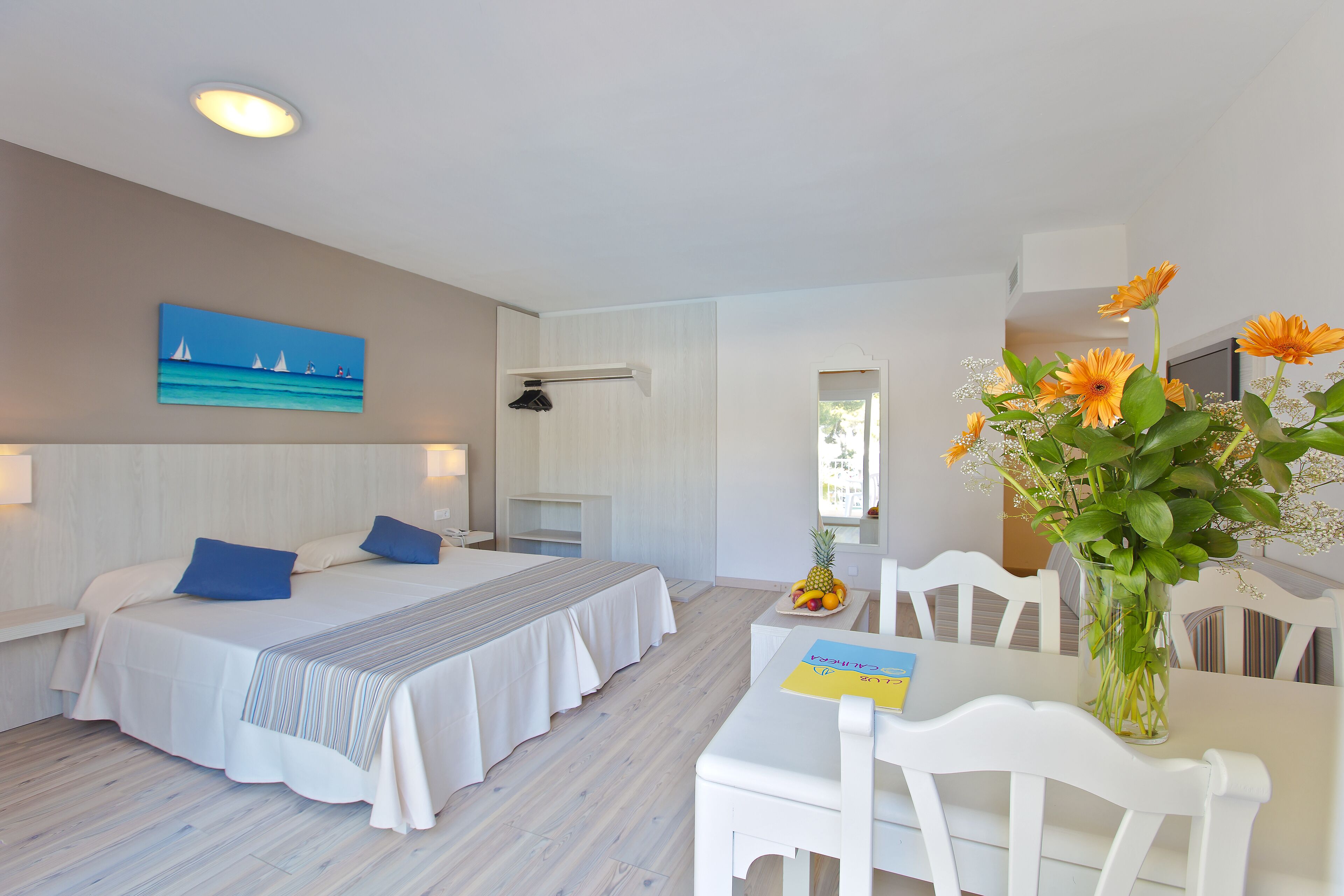 Habitación de hotel con cama, escritorio y silla. Se observa un cuadro en la pared y un florero con flores en el escritorio.