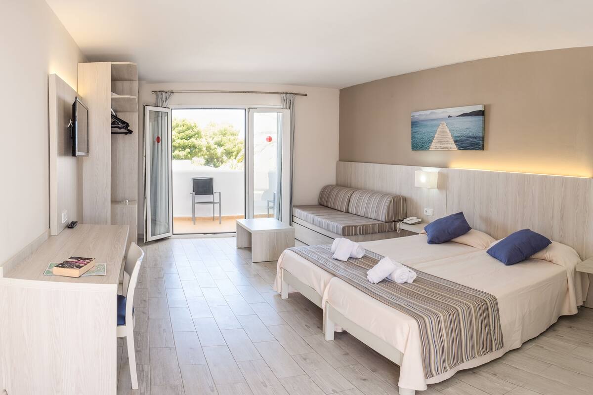 A hotel room with a bed, a desk, a chair, a small table, and a sliding glass door leading to an outdoor area.