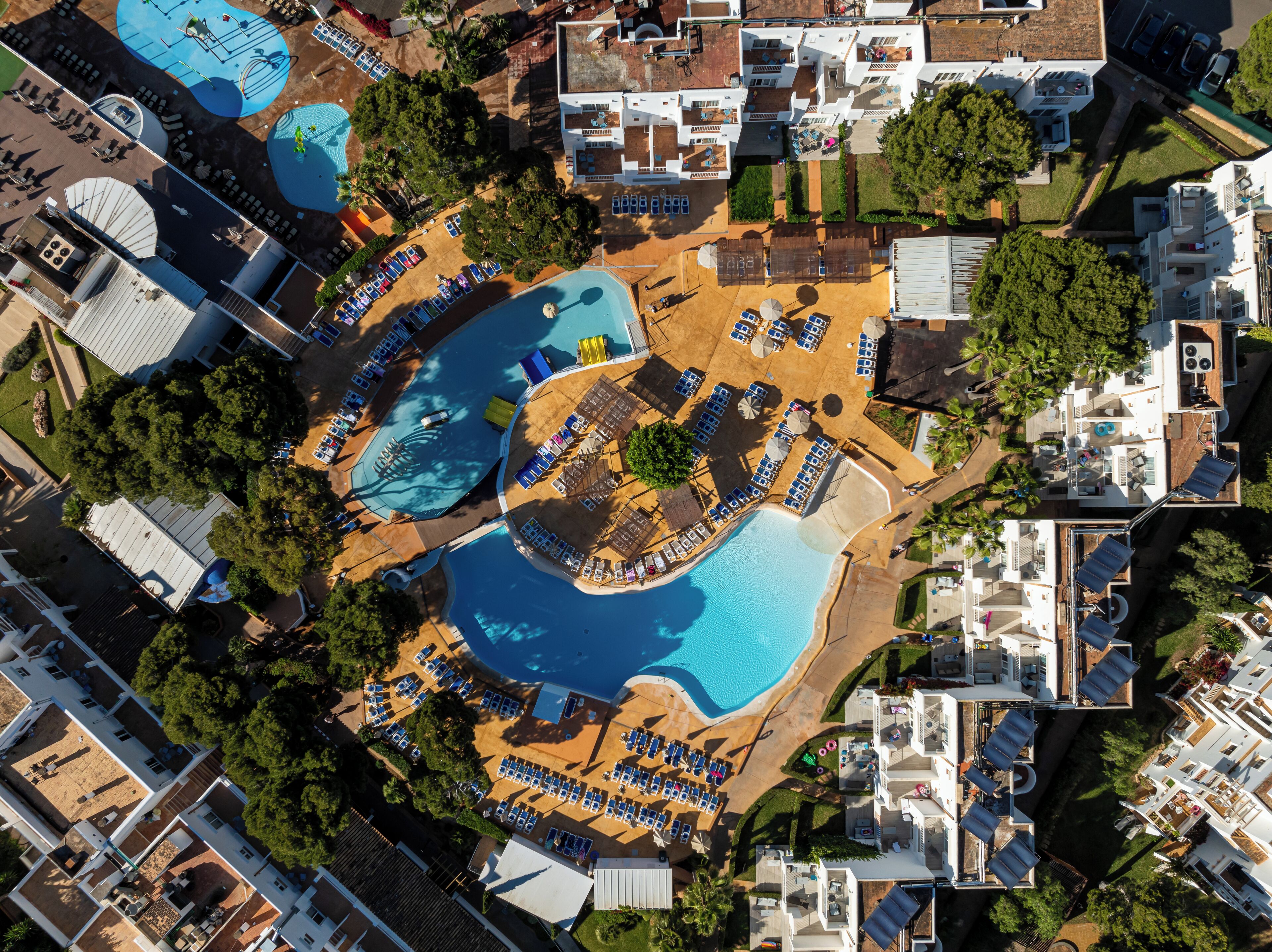 Vista aérea de un resort con dos piscinas grandes, tumbonas y una pequeña zona de playa.