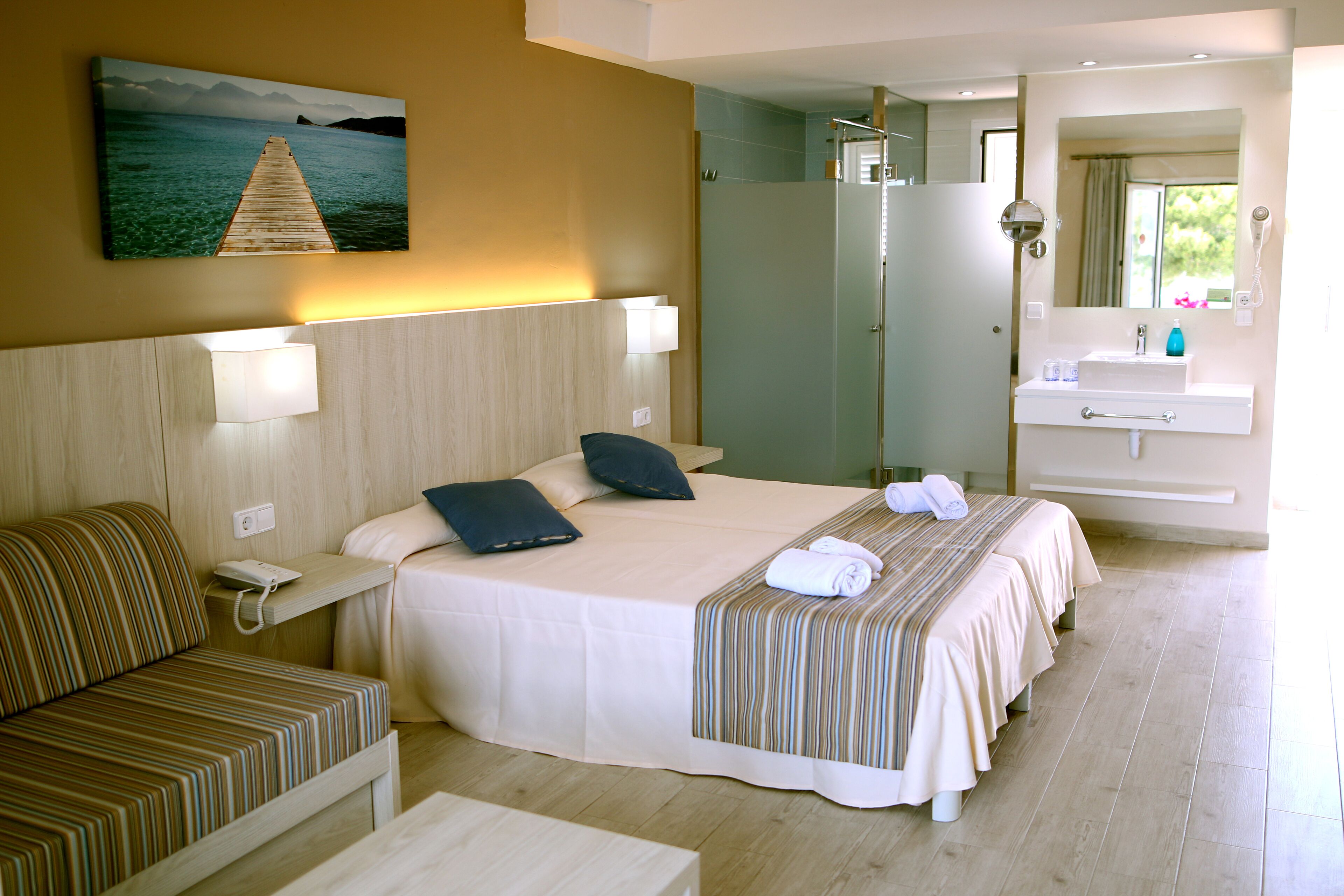 Una habitación de hotel con una cama, mesita de noche, lámpara y un cuadro de un muelle.