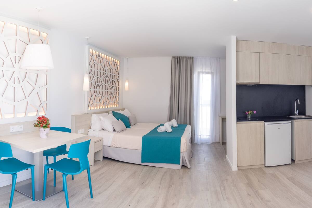 A modern hotel room with a bed, a desk with chairs, a kitchenette, and a decorative wall panel.