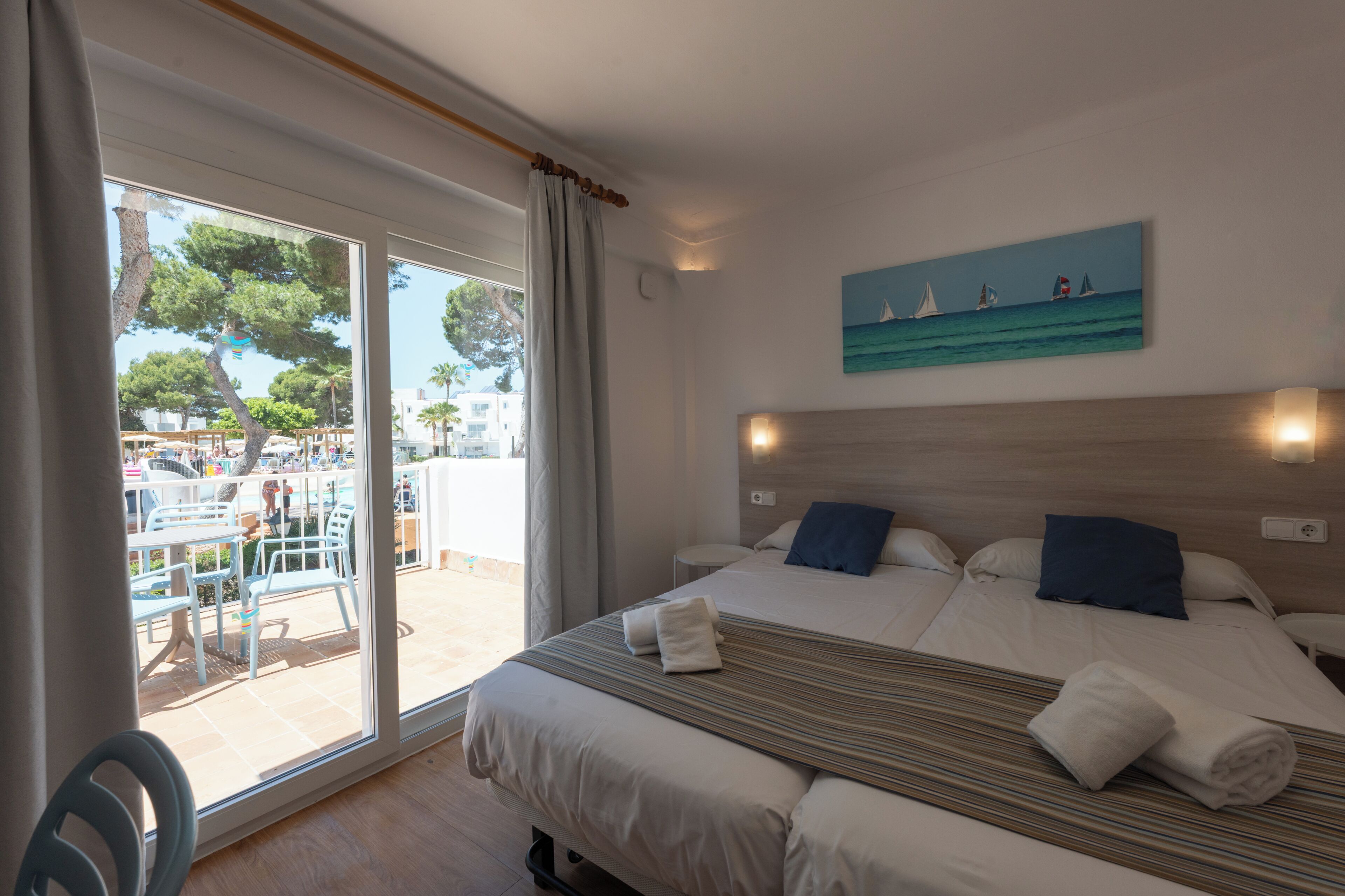 A hotel room with a bed, a painting of sailboats, and a view of an outdoor pool area.