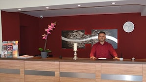 A reception desk with a man standing behind it, a clock on the wall, and a floral arrangement on the left. - Altstadthotel Harburg (Hamburg)